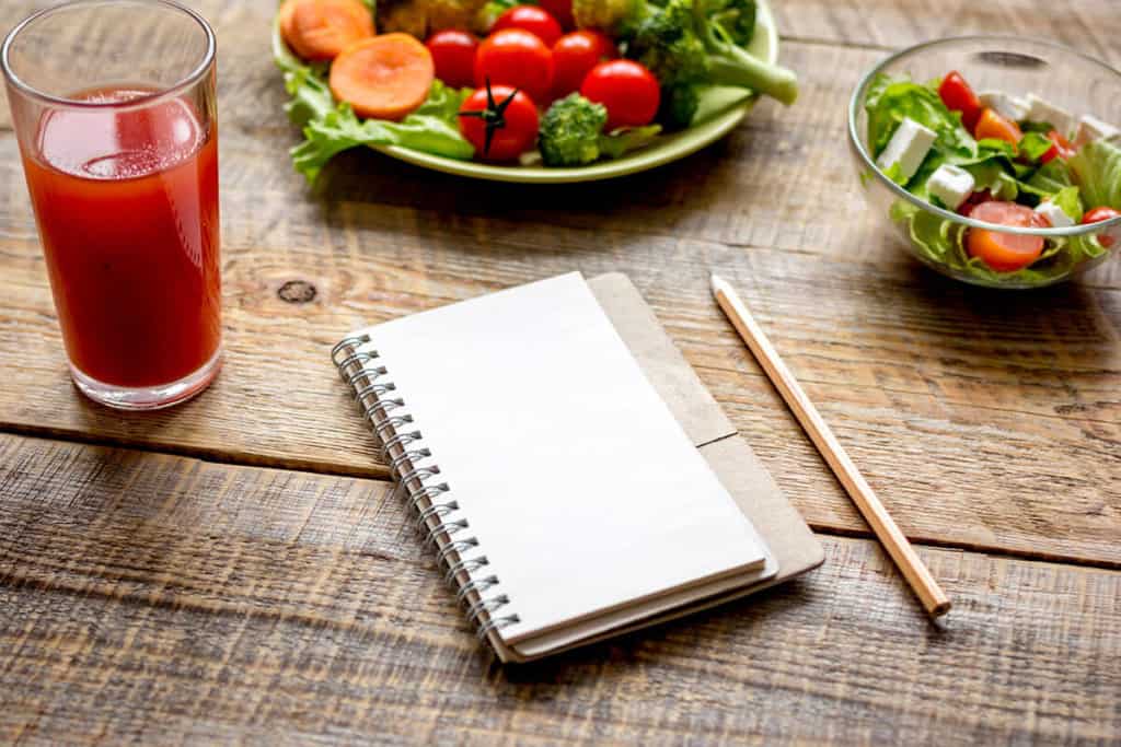 notebook with a pencil placed beside it, accompanied by a bowl of greens and a glass of juice, suggesting a balanced weight loss diet
