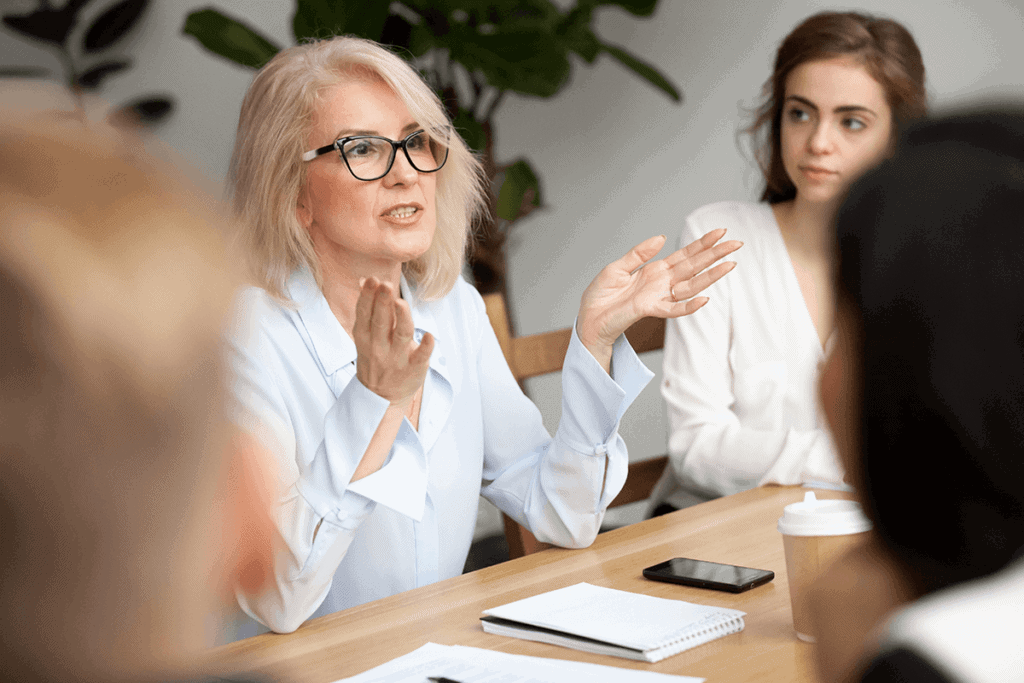 Woman sitting in an office meeting room, raising her hands while speaking to others who are listening.