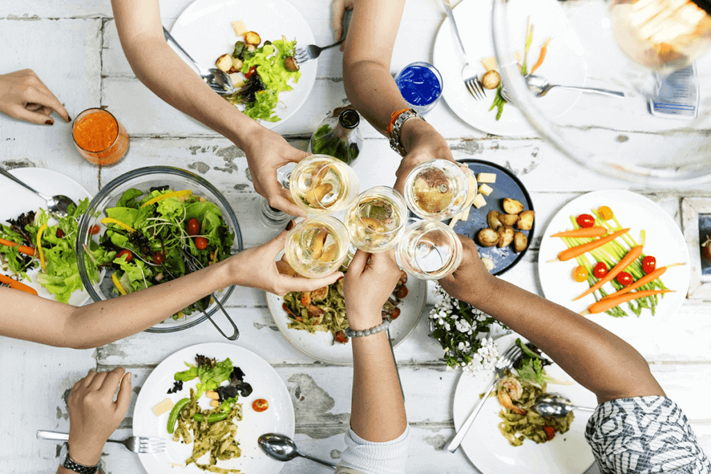 Five people holding up drinks over plates of healthy looking foods at a social picnic.