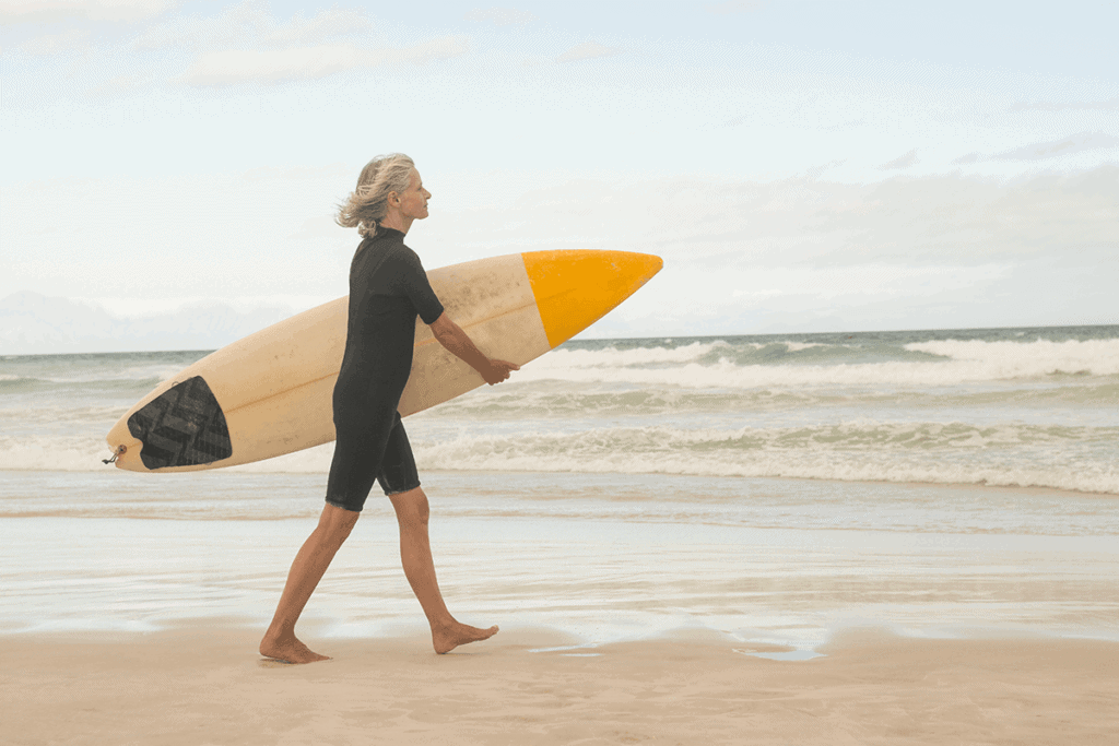 older woman walking on the beach next to the ocean carrying a surfboard